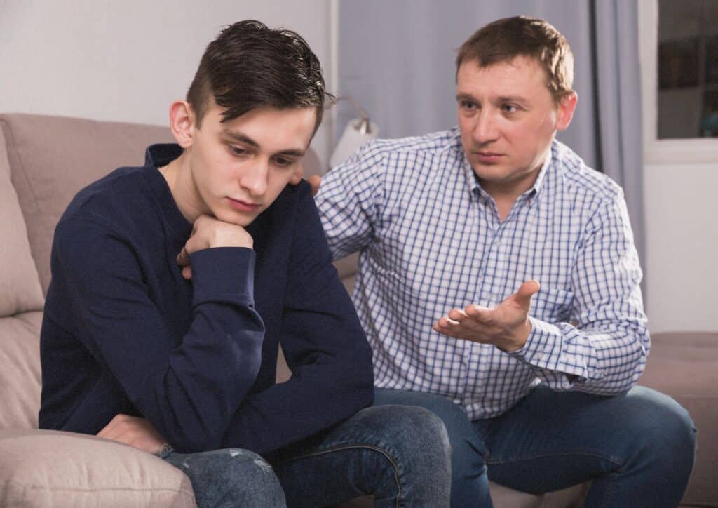 Pare assegut al costat del seu fill adolescent intentant parlar amb ell mentre el noi evita el contacte visual i mostra distància emocional, representant les dificultats de comunicació habituals durant l’adolescència.